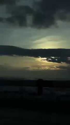 The video shows a landscape in winter at dusk. The foreground is obscured by a dark silhouette, slowly revealing a snow-covered field. As the camera pans, trees and the frame of a small building come into view, all dusted with snow. Above, the sky is a blend of blue and grey, with dark clouds partially obscuring the setting sun. The overall mood is somber and still.