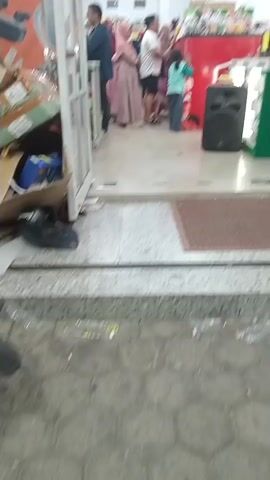 The video shows an interior view of a store, likely a supermarket or electronics shop, looking towards the entrance. Several people are visible, including a man in a dark shirt and jeans standing near the doorway, a woman in a pink hijab and long dress, a man in a white t-shirt and shorts, and a young girl in a teal and white outfit. A large black speaker is placed on the floor inside the store. Displays of products are visible on shelves in the background. The floor inside the store appears to be tiled, with a textured mat near the entrance. The exterior area in front of the store is paved with cobblestones. The lighting suggests it is daytime.