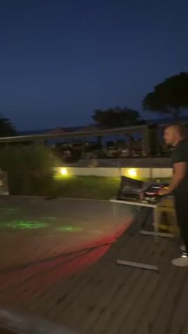 At night, people are gathered on a wooden deck outdoors. In the foreground, a DJ setup with a laptop and speakers is visible on a table. Green and red laser lights are projected onto the deck. Three people are standing near the center, two women and one man, appearing to be talking. Behind them, several other people are seated at tables under umbrellas, presumably at a restaurant or bar. The scene is illuminated by artificial lights and the dark sky, suggesting it is late evening or early night. The environment appears to be near a body of water, possibly a beach or coast, as indicated by the dark horizon.