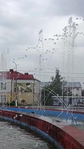 The video shows a public fountain with multiple jets of water spraying upwards against a cloudy sky. In the background, there are several buildings, including one with a red roof and a large banner featuring a portrait. There are also streetlights and trees visible. The fountain has a red and blue edge, and the water is choppy. A few indistinct figures can be seen on a sidewalk in the distance.