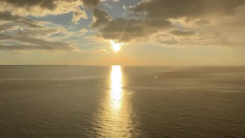 A wide shot shows a panoramic view of the ocean at sunset. The sun is low on the horizon, casting a bright, shimmering reflection across the water. Clouds are scattered across the sky, with rays of sunlight peeking through. In the distance, a few ships can be seen on the horizon. On the right side of the frame, a large cargo ship is docked at a port, with its deck and cargo visible. The overall scene is one of serene beauty, with the warm colors of the sunset creating a peaceful atmosphere. There are no people visible in the frame. The video appears to be shot from a high vantage point, possibly from another ship or a drone.