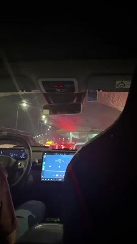 The video shows the interior of a car at night, looking forward through the windshield. The driver's side of the steering wheel is visible on the left. A large touchscreen display is in the center of the dashboard, showing a music interface. Outside, traffic lights are visible, with red and white lights illuminating the road ahead, suggesting a tunnel or underpass with traffic. The interior of the car is dark, with some ambient light reflecting off the windshield. The perspective is from the back seat looking towards the front.
