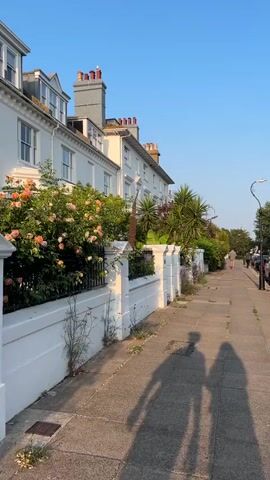 The video captures a street scene with white buildings and a sidewalk under a clear blue sky. The sun casts long shadows of two people walking towards the camera. The buildings appear to be townhouses, with ornate chimneys and windows. Lush green trees and flowering bushes are visible in front of the houses, separated from the sidewalk by a white fence and wall. A streetlamp stands on the right side of the sidewalk, and cars are parked along the street in the distance. The overall impression is one of a pleasant, sunny day in a residential area.