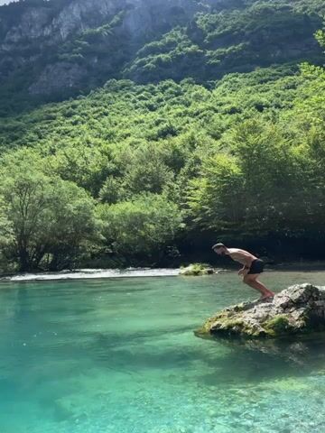 A person dives into a turquoise body of water from a rocky outcrop. The person is wearing black swim trunks. The water is clear and the bottom is visible. Lush green trees and a steep, rocky mountain form the background. The sky is partly cloudy.