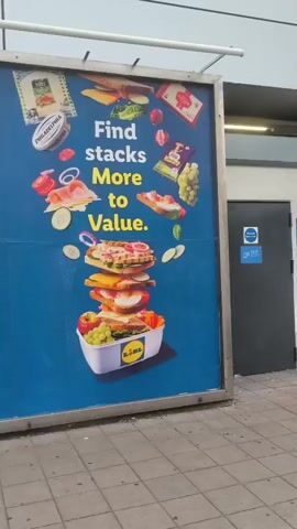 This is a medium shot video of a large blue billboard advertising Lidl. The billboard is divided into three sections, each featuring a different slogan and corresponding imagery of grocery items. The left section shows a cooler filled with frozen foods, with the text "Find freezer loads More to Value." The middle section displays a tall, stacked sandwich and lunchbox, with the text "Find stacks More to Value." The right section features a grocery bag overflowing with food items, accompanied by the text "Find bags More to Value." The billboard is located outdoors, with a sidewalk in the foreground and a building in the background. The lighting suggests it is daytime. There are no people visible in the video.