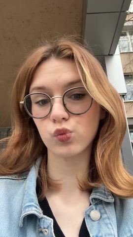 A young woman with light-reddish brown shoulder-length hair, wearing round glasses, a light blue denim jacket, and a black top, is looking directly at the camera. She briefly puckers her lips in a kissing gesture. The background includes a building with a light-colored facade and windows, and part of a roof or overhang is visible above her. Street sounds can be heard.