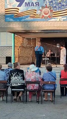 The video shows an outdoor event in front of a brick building, possibly a community center or school. A banner hangs above the stage, featuring the text "9 MAY" in large numbers, accompanied by images of doves, ribbon, and a military medal. On the stage, a man in a blue shirt and tie stands at a podium, speaking into a microphone. To his right, a person sits at a keyboard, and another person in a white garment stands next to them. In front of the stage, a group of people, mostly women, are seated on chairs, facing the stage. The lighting suggests it is daytime, with sunlight illuminating the scene. The overall atmosphere appears to be a public gathering, possibly a commemoration or celebration.