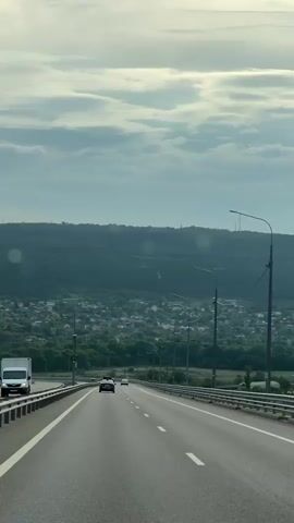 A camera within a moving vehicle records the view from the front passenger seat, panning right to reveal a burgundy SUV overtaking the camera car on a multi-lane highway. The highway is elevated, bordered by guardrails and light poles. In the distance, a town is nestled among rolling, green hills beneath a cloudy sky. The highway itself is sparsely populated with vehicles. As the SUV passes, the camera’s view focuses on the distant town and hills before being partially obstructed by the SUV's roof.