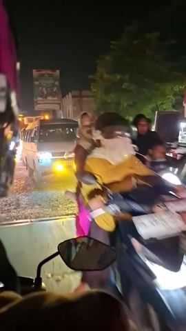 The video appears to be shot from the perspective of someone riding a motorcycle at night. The road is illuminated by streetlights and vehicle headlights. The main focus is on a white van with yellow auxiliary lights and a truck with a red and black advertisement on its side. Both vehicles are driving in the same direction as the motorcycle. Several other motorcycles can be seen briefly, as well as a couple of buildings in the background. The road surface looks uneven and somewhat gravelly. The person filming is stationary at times and slowly progressing with traffic at other times.