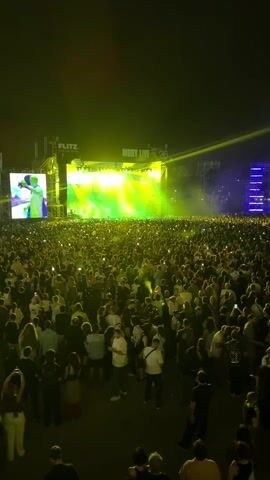 The video shows a large outdoor concert at night. A stage with bright green and blue lights is visible in the background, with a crowd of people in front of it. Many people are holding up their phones, likely recording or taking pictures. The overall atmosphere appears to be energetic and lively, typical of a music festival or concert event.