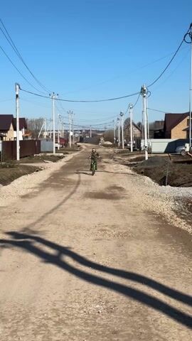 A child is riding a bicycle down a dirt road on a bright, sunny day. The child is wearing a jacket and appears to be the only person visible on the road. The road is surrounded by houses with fences and utility poles run along the side of the road. The sky is clear blue. The camera is stationary, capturing the child as they approach.