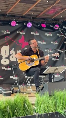 A man is playing an acoustic guitar and singing on a stage. He is wearing glasses, a striped shirt, and black pants. The stage has a black backdrop with a pink butterfly and the words "BAKgarden" repeated. There are pink stage lights above him. In front of the stage, there is a barrier of tall green grass. The man is sitting on a folding chair.