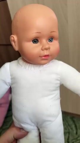 The video shows a close-up of a baby doll with a bald head and blue eyes. The doll is wearing a white onesie. It is held by a person's hands, the skin tone of which is fair. The doll is positioned in front of a wooden background. The lighting suggests it is daytime, and the setting appears to be indoors. The doll's expression is neutral, with rosy cheeks. The scene is static, with a slight blur as the doll is moved. No visible text is present. The location context provided is Почтовское сельское поселение, Ukraine.