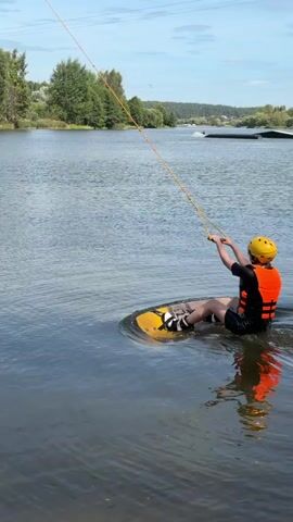 A person in a yellow helmet and orange life vest is wakeboarding on a lake. They are being pulled by a rope attached to a tow system. The person is wearing dark shorts and a black t-shirt. The wakeboard is yellow and white. The water is rippling behind the wakeboard. Trees and a blue sky with some clouds are visible in the background.