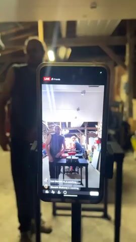 The video opens with a smartphone on a stand, displaying a live stream of an arm-wrestling match. "LIVE" and "Friends" are visible at the top of the phone screen, and "Rhamde (Rehame) is watching" appears as a comment. A man in a dark sleeveless shirt stands in the foreground, out of focus.

The scene then shifts to two men engaged in an arm-wrestling match at a dedicated arm-wrestling table with red and black pads. The man on the left wears a red sleeveless shirt and a tan baseball cap, with a focused expression. The man on the right wears a grey sleeveless shirt and a red wrist wrap, also showing intense concentration. Both men are holding onto each other's hands, locked in a struggle. A green strap with "MEE" visible is on the table next to the man in the red shirt.

The setting appears to be an indoor, rustic space, possibly a garage or workshop, with exposed wooden beams on the ceiling and artificial lighting. A light-colored, possibly Japanese-style, folding screen is visible behind the arm-wrestlers. Other equipment and clutter can be seen in the background, suggesting a functional, rather than polished, environment. The overall ambiance suggests an evening or night setting due to the reliance on artificial light.