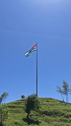 A video shows the flag of Abkhazia flying on a tall flagpole atop a grassy hill. The flag is green and white striped with a red canton featuring a white open hand and seven white stars. The flag gently waves in the breeze against a clear, bright blue sky. A few small trees are visible on the hill. The sun is shining brightly, indicating daytime.