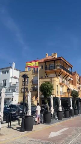 The video shows a bright, sunny day in what appears to be a coastal town in Spain. A Spanish flag flies prominently from a tall flagpole atop a tan-colored building with black balconies. The building has "CASA" visible on its front. The area around the building is a pedestrian zone with outdoor seating for a restaurant or cafe, featuring white umbrellas, tables, and barrel-shaped planters.  A young man in a white shirt and shorts stands near the front of the building, looking at something out of frame.  Several cars, including a dark SUV, are parked along the street.  Across the street is a white building with a tower. The sky is clear and blue.