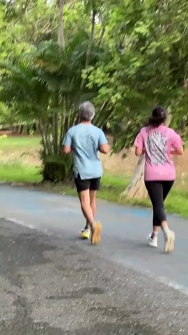 Two individuals are seen from behind jogging on a paved path. The person in the foreground on the left is wearing a light blue t-shirt, black shorts, and yellow shoes. The person on the right is wearing a pink t-shirt with a graphic design on the back, black leggings, and white shoes. Both are running away from the camera. The path is bordered by lush green trees and foliage, suggesting an outdoor setting like a park or trail. The ground appears to be slightly wet, possibly from recent rain.