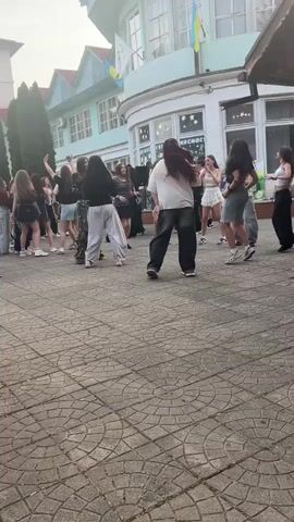 A group of teenagers are dancing outside a building. The building appears to be a school with a light blue facade and a rounded corner. There is a Ukrainian flag hanging from the building. The ground is paved with interlocking bricks. The teenagers are dressed casually in t-shirts, shorts, and pants. Some are dancing in pairs, while others are dancing individually. The overall atmosphere appears to be festive and energetic.