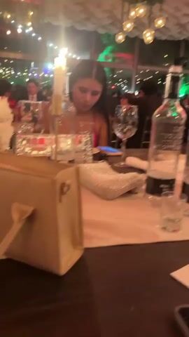 The video shows a dimly lit indoor event, possibly a wedding reception or banquet. People are seated at tables adorned with white floral centerpieces and lit candles. The lighting is warm and ambient, with fairy lights visible in the background, suggesting it is evening. In the foreground, a woman in a pink top is looking down at her phone. To her left, a purse rests on the table. Further to the left, another woman is partially visible, also at a table setting. In the middle ground, a man in a suit is looking down, and to his right, another woman appears to be speaking. The overall atmosphere is one of a social gathering.