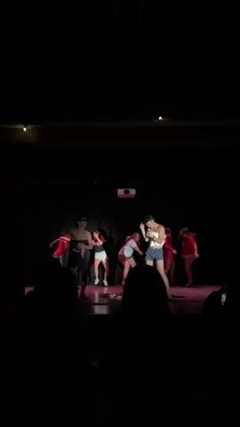 A group of seven people are on a stage in a dark room. The person on the far left is wearing a black strapless top, sunglasses, and shorts. They are holding a pink cloth. The person in the center is wearing a light-colored top and shorts and is holding a microphone. The other five people are wearing red tops and shorts and are dancing or posing. There is a projector mounted on the ceiling. The stage is lit with pink and white lights.