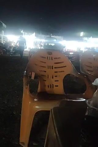At night, the video shows an outdoor cafe or market with several tables and chairs under green umbrellas. Bright lights illuminate the area, revealing people sitting at the tables and moving around. The camera focuses on a table with a cup of something and plastic chairs, moving slightly and providing a view of the scene. The ground appears to be a dark, gravelly surface.
