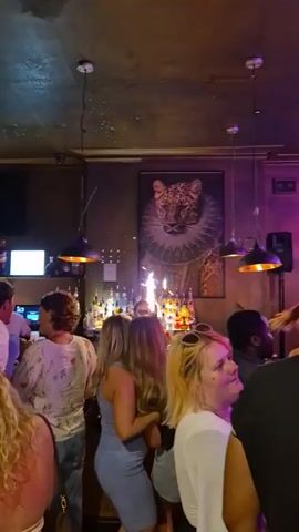 Okay, here is a brief, matter-of-fact description of the video:

The video shows a crowded bar with people standing around the bar counter. Some are talking, and at one point, sparklers are lit behind the bar near the liquor bottles. The bar is decorated with hanging lights and a framed picture of a leopard in a ruff collar. Several people are looking toward the bar where the sparklers are.