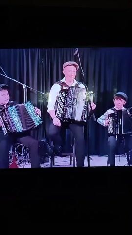 Three people are seated on chairs on a stage, playing accordions. The person in the center, an adult male wearing a white shirt, dark pants, and a flat cap, is playing a large accordion. To his left, a young person, likely a child or teenager, wearing dark clothing, is playing a smaller accordion. To the right of the central figure, another young person, also wearing dark clothing, is playing a similar-sized accordion. The background consists of dark curtains with some faint lighting or decorations, and microphones are visible. The overall scene appears to be a performance.