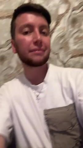 A young man with short dark hair and a beard, wearing a white t-shirt with a gray pocket and a silver necklace with a cross, is looking at the camera against a marble-patterned wall. He smiles, talks, and makes hand gestures, occasionally touching his face and hair.