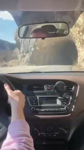 A driver in a light-colored sweatshirt is driving a car through a natural rock tunnel. The driver's left hand is on the steering wheel. The interior of the car is visible, showing a brown dashboard, a radio, and a phone mount. The rearview mirror reflects the driver's head. The road surface is visible through the windshield. The tunnel walls appear rocky and natural.