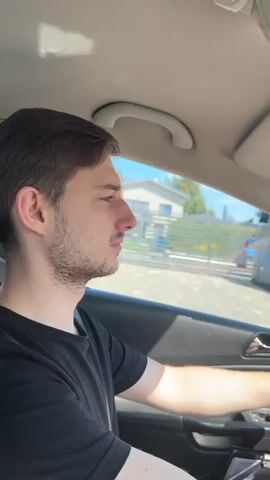 The video shows a young man driving a car. The car interior is visible, including the dashboard, steering wheel, and rearview mirror. The man is wearing a black t-shirt and looking ahead. Outside the car, trees and buildings are visible, suggesting a suburban or rural setting. The sun is shining, and the sky is blue. The video appears to be filmed from inside the car, with some shots focused on the driver and others on the road ahead.