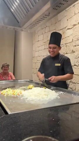 A male chef in a black uniform and chef's hat smiles as he prepares food on a large metal griddle. He holds a spatula in his right hand and a metal container in his left, from which he is adding ingredients to a large pile of white rice. To the left of the rice is a pile of chopped vegetables, including carrots, onions, and peas. A woman with short gray hair and a floral top sits at the griddle in the background, looking at the chef. The griddle is in a restaurant kitchen with a stainless steel hood above it and a stone wall behind the chef.