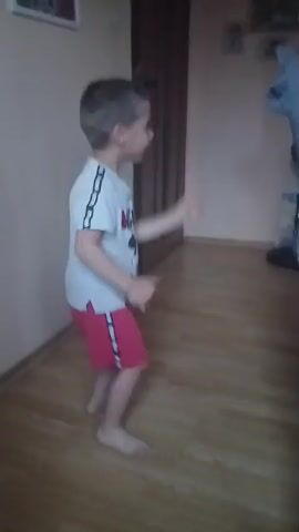 A young boy stands indoors on a wooden floor, wearing a white t-shirt with a Mickey Mouse design and red shorts with white stripes. He looks to his right, moves his arms in front of him and then turns to face the camera. The room has light-colored walls. A brown door is visible in the background, along with some small pictures on the wall. There is a chair and a bag near the wall as well.