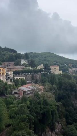 The video pans across a hillside village with multi-colored buildings nestled amongst lush green trees. The sky is overcast with dark, dramatic clouds. In the foreground, a decorative lamppost and a stone railing are visible. Flags are displayed on poles, adding a touch of color to the scene. The architecture suggests an Italian setting, with terracotta roof tiles and stucco facades. The overall atmosphere is one of a slightly moody, historic European town.