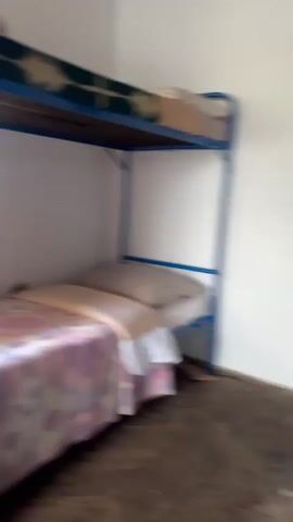 The video shows the interior of a room with bunk beds. The lower bunk is made with a patterned comforter and pillows. A window to the left of the bed shows a glimpse of the outdoors, and a curtain partially obscures the view. The camera then moves closer to the window, revealing a mountainous landscape with snow-capped peaks and a lake. Wooden structures and a fence are visible in the foreground, suggesting an outdoor area. The lighting is bright, indicating daytime.