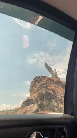 The video shows a large eagle statue perched atop a rocky cliff face. The statue appears to be made of metal and is facing to the right with its wings outstretched. The cliff is dry and rocky, with sparse vegetation. Below the cliff, a road is visible with a white car driving on it. The sky is blue with some scattered clouds. The video is filmed from inside a vehicle, with the edge of a car window frame visible in the foreground.