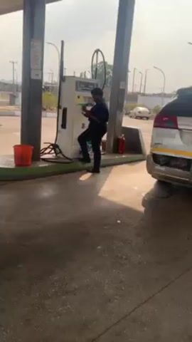The video shows a man standing next to a gas pump at a gas station. He is holding a phone and appears to be looking at it. A white minivan is parked next to the pump. Another man in blue pants stands behind the van, also looking at his phone. The gas station has a green and white color scheme. There is a fire extinguisher next to the gas pump. The ground is wet and shiny.