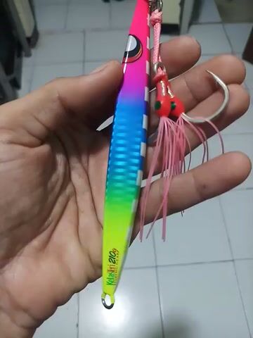Here's a concise description of the video:

A person's hand holds a colorful fishing lure. The lure is long and thin, colored with stripes of pink, white, rainbow gradients, and yellow-green. It has a hook attached at the top with pink strands. The lure has the word "Kotosen 210g" printed on it in yellow letters. The background is a tiled floor.
