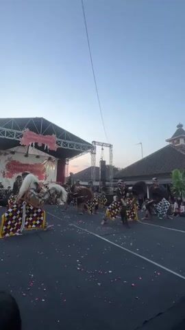 A group of performers, some wearing horse-like masks and elaborate costumes, are engaged in a synchronized dance on a paved outdoor area. The performers' attire features colorful, checkered patterns. In the background, a stage is set up with banners and lighting. There are spectators seated and standing to the right of the performance area. The sky is clear and appears to be during either sunrise or sunset. The overall scene depicts a cultural performance or festival.