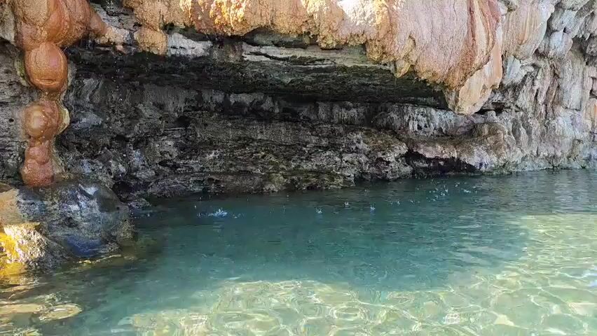 The media file depicts a natural scene at Loutra Edipsou, Greece. The foreground is dominated by clear, turquoise water, with ripples indicating subtle movement or possibly drips falling from the overhang above. The water appears shallow near the shore, allowing the sandy or rocky bottom to be visible in places.

The focal point is a large, overhanging rock formation, heavily encrusted with mineral deposits. These deposits are predominantly in shades of orange, brown, and white, giving the rock a textured, almost sculpted appearance. They hang down in stalactite-like formations, some smooth and rounded, others rough and irregular. Beneath the overhang, the rock face is dark and layered, suggesting geological strata. Water appears to be seeping or dripping from various points on the overhang, contributing to the mineral formations and the ripples in the water below.

The background shows a continuation of the rock face, leading upwards and to the right. The sky is a clear, bright blue, suggesting fair weather and likely daytime. There are hints of greenery on a distant hillside in the far right of the frame. The overall impression is one of natural beauty and geological wonder, likely a popular tourist or bathing spot given the clear water and accessible formation. No people or specific objects are clearly identifiable, and there is no visible text. The scene evokes a sense of tranquility and the raw power of nature.