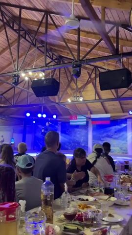A video shows a man playing an accordion on a stage in front of an audience. The stage has blue lighting and two flags, one red, white, and blue and another with green and white stripes. In front of the stage, people are seated at tables with food and drinks. The room has a wooden ceiling with exposed beams and a fan.