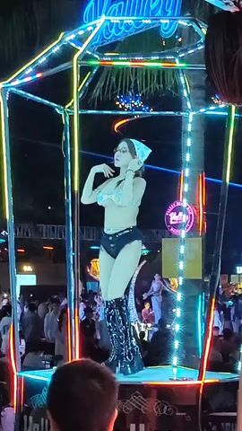 Here is a brief description of the video:

The video shows a woman dancing on a platform in an outdoor setting. The platform is enclosed by a cube-shaped structure outlined with neon lights. The woman is wearing a bikini top, shorts, and high boots. She also has a headscarf and glasses. A crowd of people is visible in the background. A sign that reads "Valley" can be seen above the dancer. The scene appears to be from a nightlife establishment.