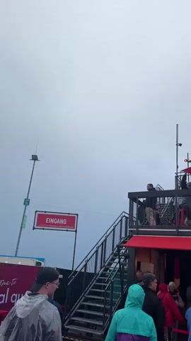 The video shows an outdoor gathering with many people under an overcast sky. A multi-level red and black structure with a balcony is on the right, featuring LED screens displaying German text and signs like "FREIKOM BAR" and "SALON." People are visible on both levels of this structure. On the left, a red sign indicates "EINGANG" (Entrance) and another "AUSGANG" (Exit). As the camera pans left, a large stage-like structure with two prominent screens comes into view. Initially blank, one screen later displays a pink crown and German text, "HEUTE REGIERT DIE K." The stage also features speaker stacks with "STAY LOUD" written on them. A banner on the ground reads "WELCOME TO real vibes." Most attendees are dressed casually, many wearing hooded jackets or rain ponchos, suggesting cool or rainy weather. People are walking, standing, and socializing on the ground, with some holding food containers.