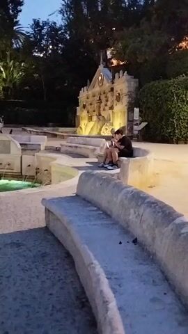 A video shows a tiered fountain in a park at dusk. A person sits on a curved stone bench facing the fountain, looking at a phone. In the background, water flows down the fountain's levels and a few other people are visible. Trees and bushes surround the fountain, and artificial lights illuminate the scene.