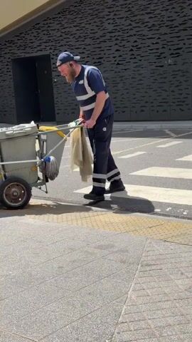 A man wearing a dark blue uniform with reflective stripes, a blue baseball cap, and a beard pushes a cleaning cart. The cart has a grey bin, a mop, and a white cloth. He is on a paved area with yellow lines painted on the asphalt. Behind him is a building with a dark patterned facade. He is walking away from the camera towards a large gate with signs.