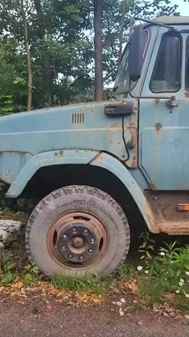 The video shows a close-up side view of a blue, rusty military truck. The truck is parked on a dirt and gravel surface with green grass and white wildflowers growing around its tires. The truck's body is weathered and shows signs of age and disuse. The cabin has two doors, and there are two red cross symbols visible on the side of the rear cargo area. The scene appears to be outdoors in a wooded or overgrown area, with trees visible in the background.
