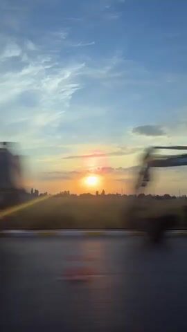 The video shows a sunset with a bright sun low in the sky, casting a warm glow. The sky is a mix of blue and orange hues, with some wispy clouds. In the foreground, there are blurred shapes of vehicles moving, suggesting the footage was taken from a moving car on a road. The road itself is visible as a dark, streaky surface with painted lines, indicating a pedestrian crossing or lane dividers. The overall scene is one of motion and natural beauty, with the sunset dominating the frame.