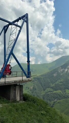 A person stands on a platform next to a large blue swing structure. The swing has a red seat and appears to be in motion. The platform overlooks a vast, green, mountainous landscape under a cloudy sky. The person raises their hand. The swing moves back and forth, carrying a rider. The rider is obscured by the swing's mechanism. The scene appears to be an outdoor amusement or adventure attraction situated in a dramatic natural environment.