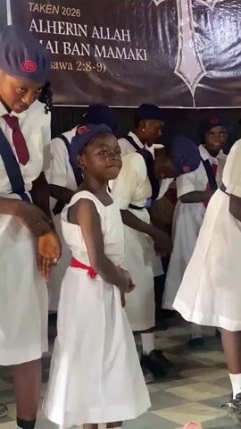 A group of young girls, all wearing similar white dresses and dark blue berets, are singing and dancing. A banner behind them features an emblem, a cross, and writing, which includes the phrase "ALHERIN ALLAH MAI BAN MAMAKI." The girls move their hands in rhythm and seem to be enjoying the performance. The camera focuses on the girl in the center as she sings. The room has a patterned floor.