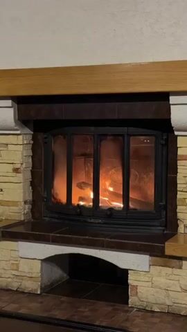 A fireplace with a glass door shows a fire burning inside. The fireplace is made of stone and has a wooden mantle. The fire is bright orange and yellow, with visible flames. The glass door is slightly dirty, obscuring some of the details within the firebox. The video is static and focuses on the fireplace.